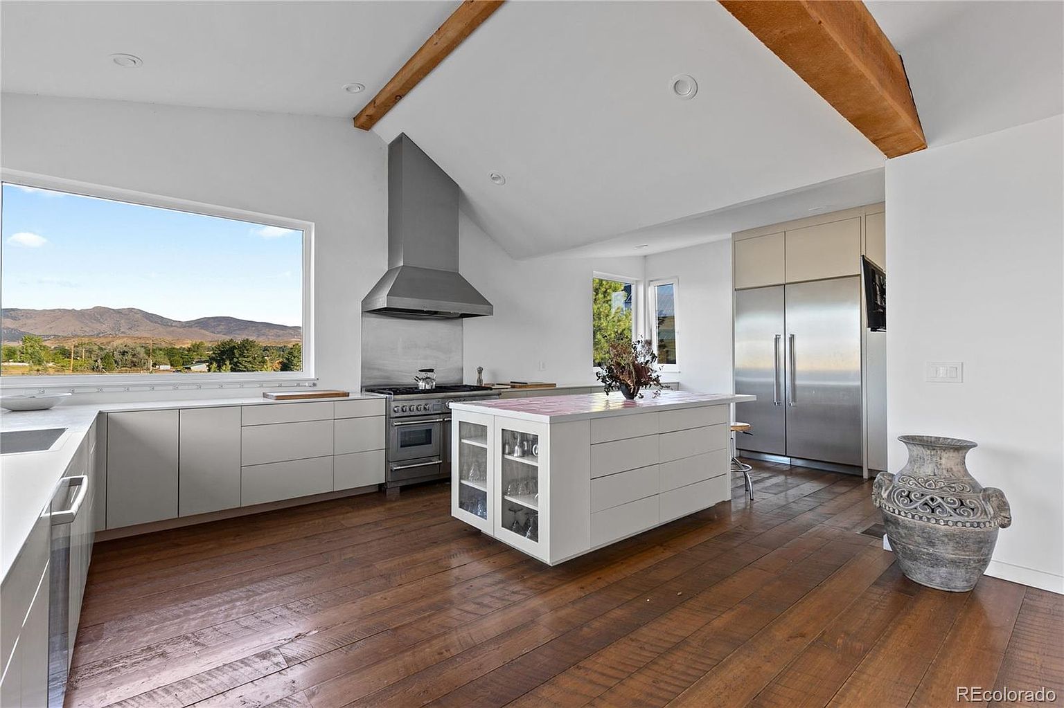 This is a bright and modern kitchen featuring sleek, light-colored cabinetry and stainless steel appliances. A large island with a contrasting countertop provides ample workspace, while a large window offers a scenic view. The hardwood flooring adds warmth to the space, and the overall design is clean and contemporary.