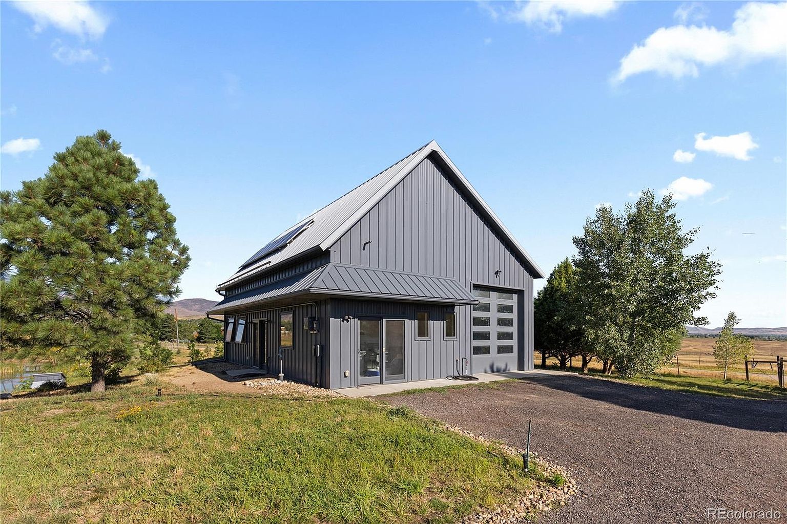 This is a front exterior view of a modern barn-style home. The house features gray vertical siding, a metal roof, and a garage door with multiple windows. The property includes a gravel driveway, a well-maintained lawn, and mature trees, creating a serene and private setting.