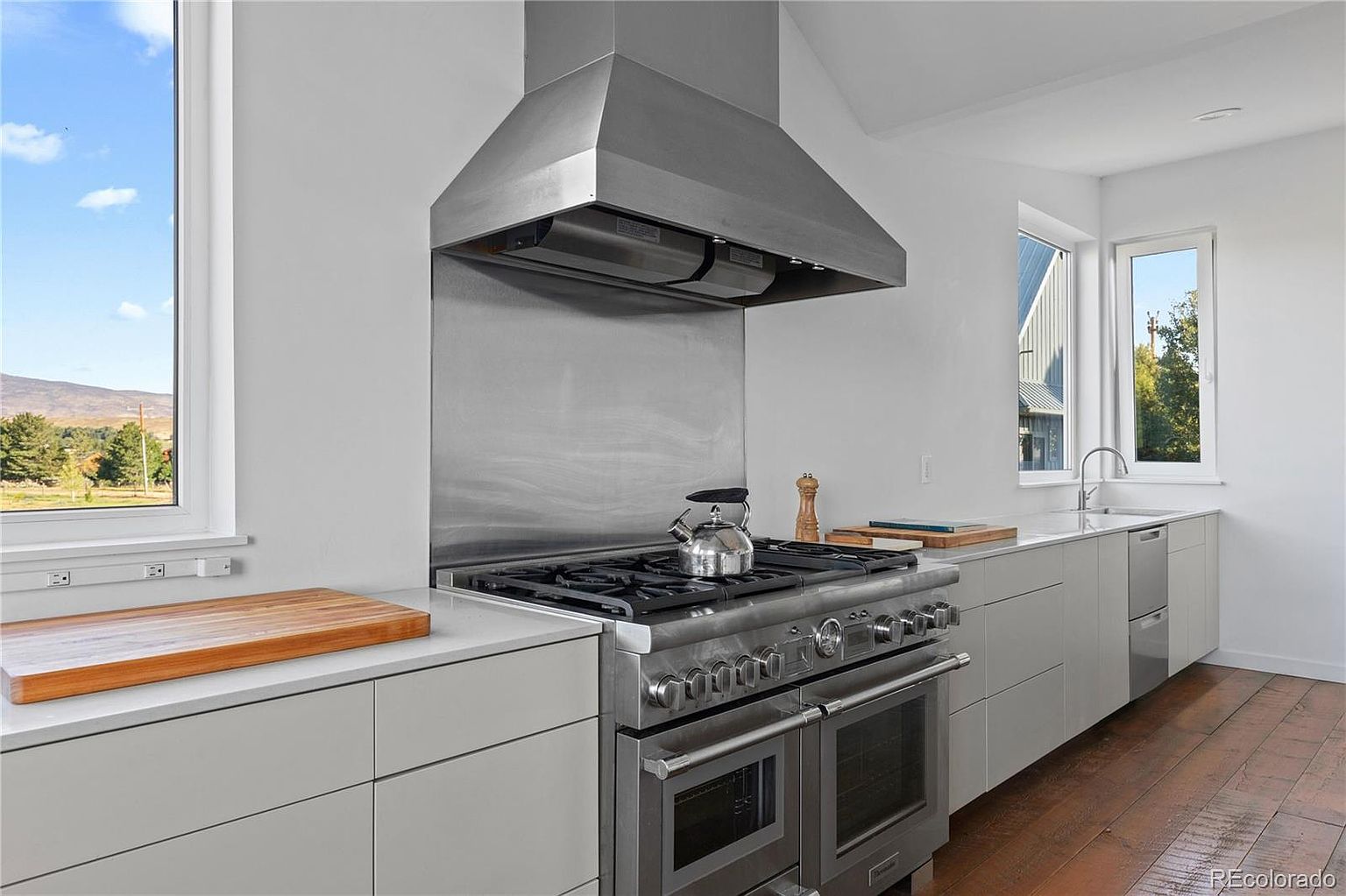 This is a modern kitchen featuring stainless steel appliances, including a large range with a hood, and light gray cabinetry. A window provides natural light and a view of the outdoors. The kitchen has a clean and minimalist design with hardwood floors adding warmth.