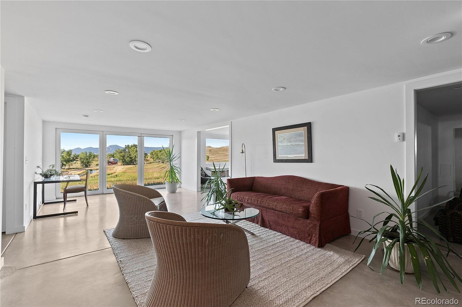 This is a bright and airy living room featuring a neutral color palette with a pop of color from the sofa. The room is furnished with two wicker chairs, a glass coffee table, and a red sofa, all placed on a light-colored rug. Large windows offer a view of the outdoors, and the space is decorated with plants, creating a welcoming and comfortable atmosphere.