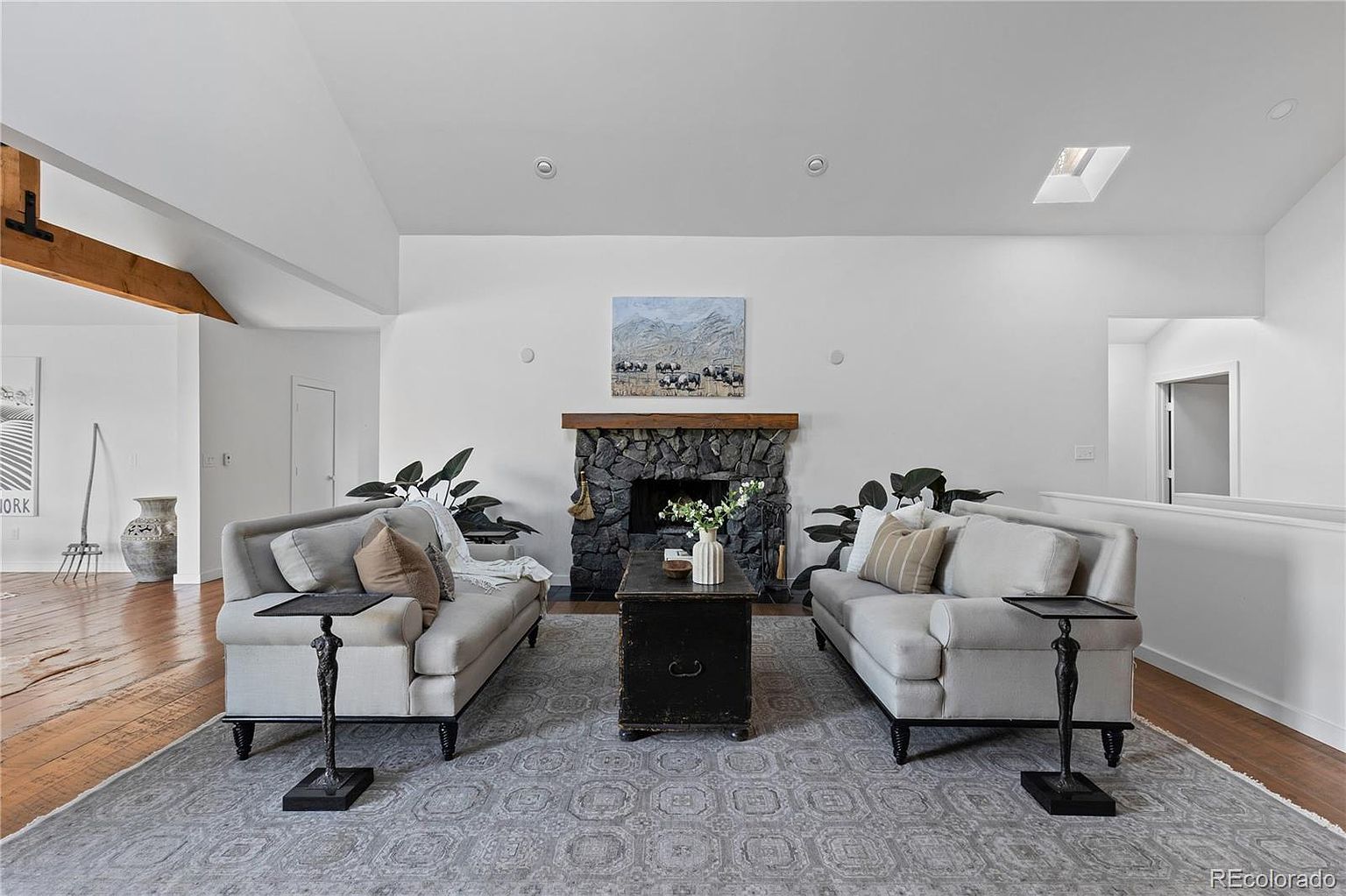 This is an interior shot of a living room featuring two light-colored sofas facing each other, flanking a dark wooden coffee table. A stone fireplace with a wooden mantel is centered between the sofas, with artwork hanging above it. The room is well-lit with natural light and features a patterned gray rug, creating a cozy and inviting atmosphere.
