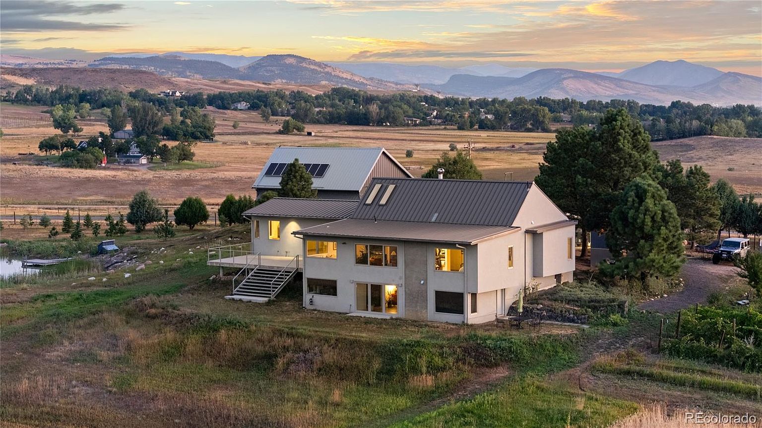 This aerial view showcases a modern home with a metal roof, complemented by a separate structure with solar panels. The property is situated on a vast, grassy landscape with rolling hills and mountains in the background, creating a serene and private setting. A small pond and mature trees add to the natural beauty of the estate.