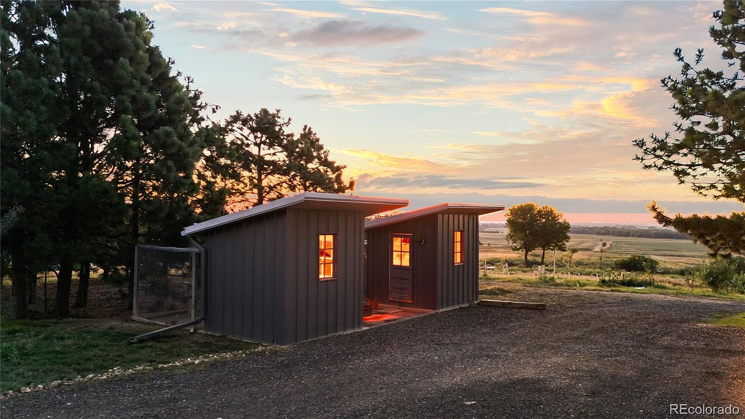 The image showcases two small, modern outbuildings, possibly chicken coops or storage sheds, set against a picturesque landscape at sunset. The structures feature dark siding and simple, clean lines, with warm light emanating from their windows. The surrounding yard is gravel, and the background reveals a scenic view of fields and a colorful sky, creating a tranquil and inviting atmosphere.