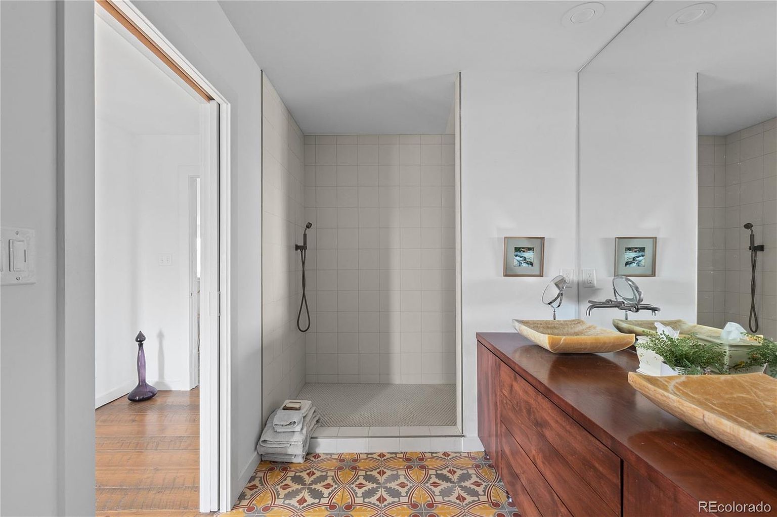 This bathroom features a unique design with patterned tile flooring and a modern shower with light gray tiling. A wooden vanity with vessel sinks adds a touch of warmth, complemented by a large mirror above. The space is well-lit and presents a clean, stylish aesthetic.