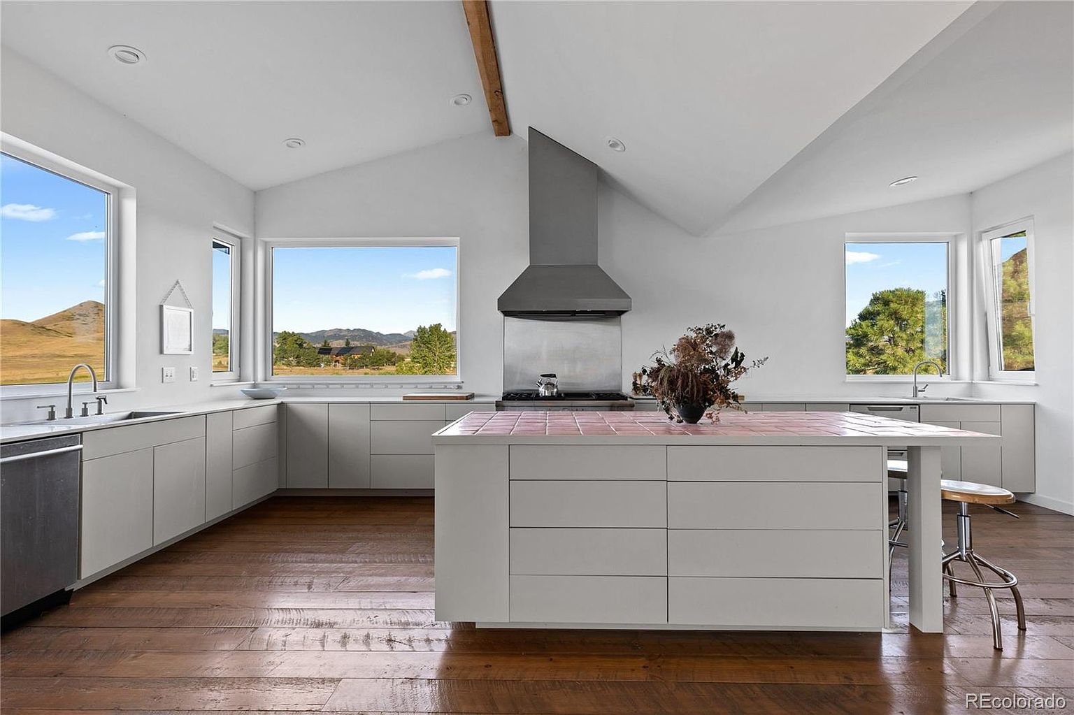 This is a bright and airy kitchen with a modern farmhouse aesthetic. The kitchen features light gray cabinetry, stainless steel appliances, and a large island with a unique pink tile countertop. The hardwood floors add warmth to the space, and large windows offer scenic views of the surrounding landscape.