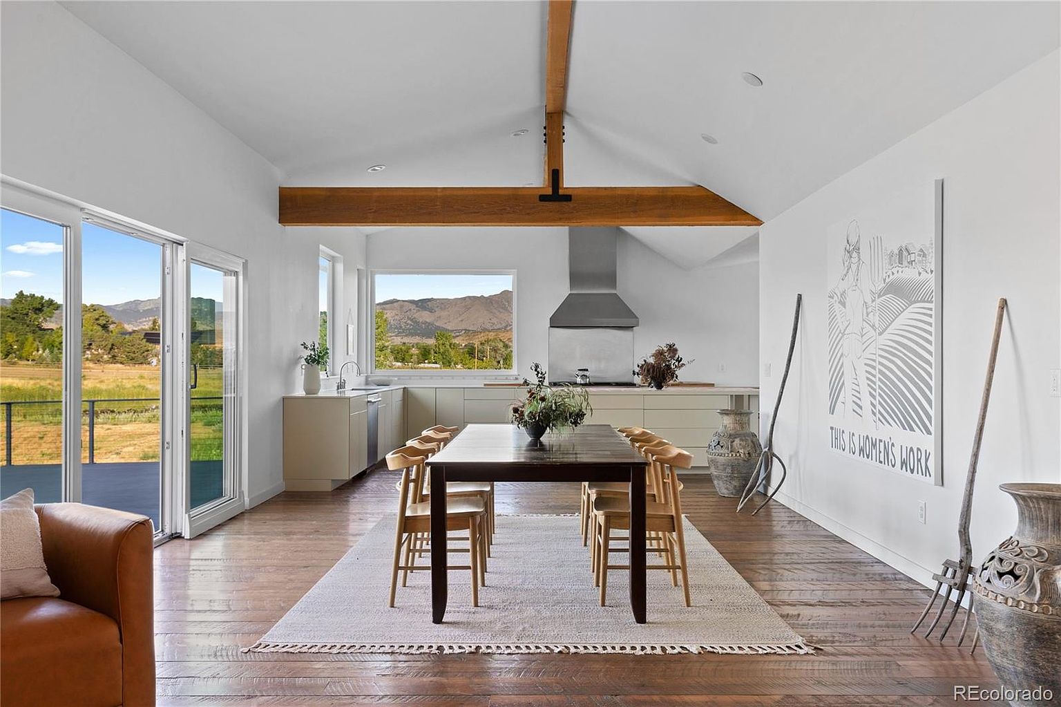 This interior shot showcases a dining area with a long wooden table surrounded by matching chairs, set upon a light-colored rug. The room features hardwood floors, white walls, and exposed wooden beams on the ceiling, creating a rustic yet modern aesthetic. Large windows and a sliding glass door offer views of the outdoor landscape, enhancing the room's bright and airy feel.