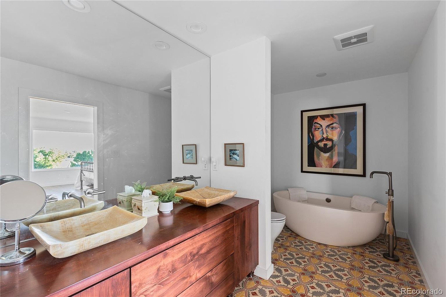 This is a primary bathroom featuring a double vanity with unique vessel sinks and modern fixtures. A large mirror spans the vanity area, reflecting natural light. The bathroom also includes a freestanding bathtub with a floor-mounted faucet, complemented by a striking piece of art and patterned tile flooring, creating a luxurious and stylish space.