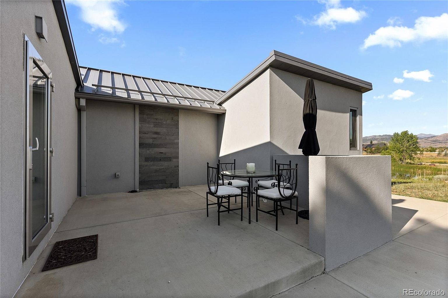This image showcases a modern patio area with a sleek, minimalist design. A round glass-topped table with four wrought iron chairs and white cushions sits on a smooth concrete surface. The patio is enclosed by a low wall and adjacent to a building with a metal roof and a unique stone accent wall, creating a private and stylish outdoor living space.