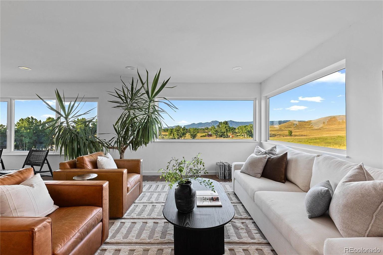 This is a bright and airy living room featuring a neutral color palette with pops of brown from the leather chairs. Large windows offer stunning views of the landscape, bringing the outdoors in. The room is furnished with a white sofa, leather armchairs, a black coffee table, and a patterned rug, creating a comfortable and stylish space.