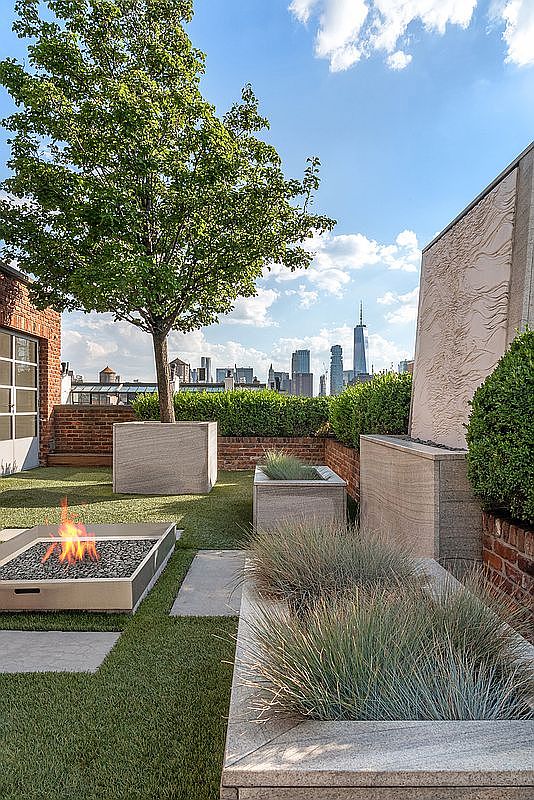 This sophisticated rooftop terrace offers a luxurious urban retreat with a stunning panoramic view of the New York City skyline, including One World Trade Center. The space is meticulously landscaped with manicured hedges, ornamental grasses in stone planters, and a mature tree, all set against a backdrop of classic brick architecture. A modern, low-profile fire pit serves as a central focal point, creating an inviting atmosphere for evening relaxation in a high-end metropolitan setting.