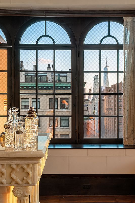 This interior shot captures a sophisticated living space featuring a classic marble console table adorned with elegant glass decanters. The focal point is a pair of large, arched windows with wire-reinforced glass that frame a stunning, iconic New York City skyline view, including the One World Trade Center. The dark wood trim and architectural detailing suggest a refined, historic, or luxury urban residence.
