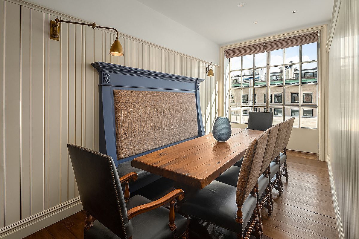 This elegant dining room features a rustic wooden table paired with a custom-built blue banquette upholstered in a patterned fabric. The space is accented by vertical striped wallpaper, sophisticated brass wall sconces, and dark leather chairs, creating a refined yet cozy atmosphere. A large window at the end of the room allows natural light to flood the space, highlighting the rich wood flooring and the sophisticated, traditional design aesthetic.