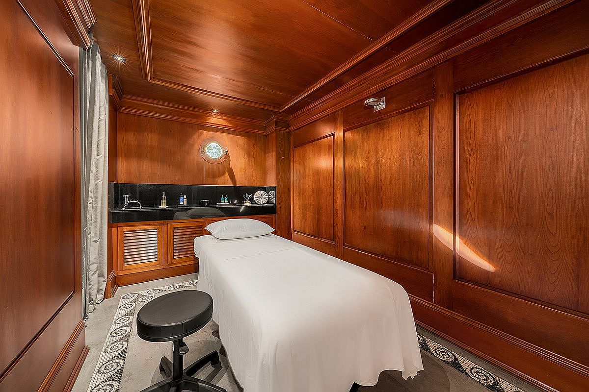 This image depicts a luxurious, wood-paneled spa or massage room featuring a professional treatment table covered in crisp white linens. The room is characterized by rich, warm-toned wood walls and ceiling, a black marble-topped vanity with a sink, and a circular porthole-style window, suggesting a nautical or high-end yacht aesthetic. A black rolling stool sits in the foreground, and the space is accented by intricate mosaic tile flooring, creating an atmosphere of refined relaxation and exclusivity.