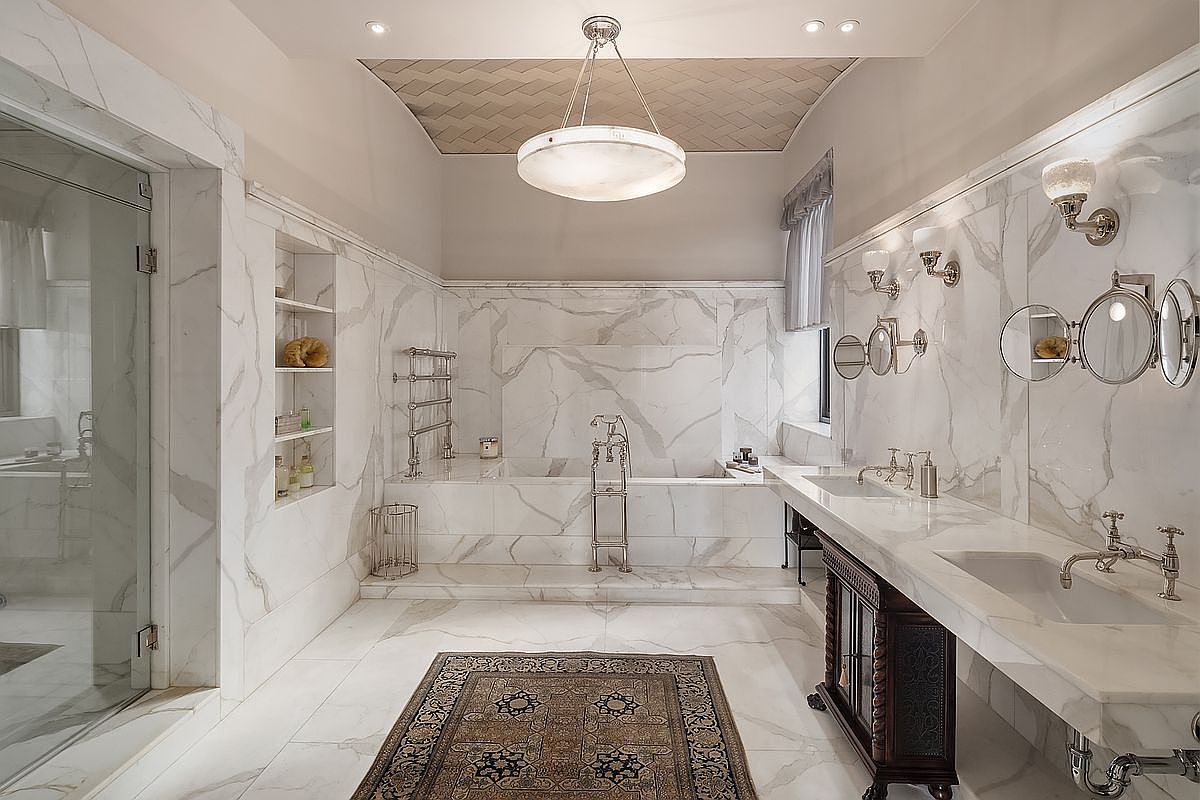 This luxurious primary bathroom features floor-to-ceiling white marble with dramatic grey veining, creating a sophisticated and opulent atmosphere. The space includes a deep soaking tub set against a marble backdrop, a double vanity with ornate dark wood cabinetry, and a glass-enclosed shower. A large, elegant chandelier hangs from a textured ceiling, while a patterned area rug adds warmth to the polished marble floor.