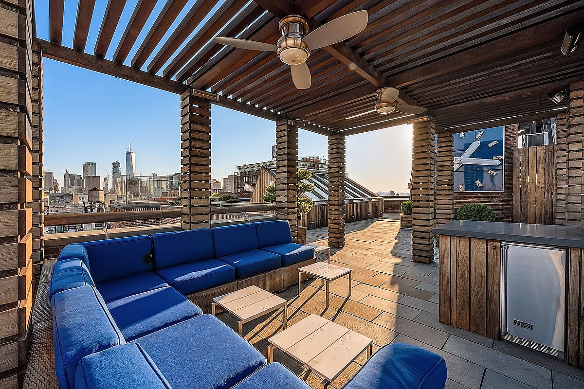 This luxurious rooftop terrace features a spacious, modular blue sectional sofa arranged under a modern wooden pergola with integrated ceiling fans. The space includes a built-in outdoor bar with a Viking refrigerator, all set against a stunning, panoramic backdrop of the New York City skyline. The combination of warm wood textures, vibrant seating, and high-end amenities creates an inviting and sophisticated urban oasis.