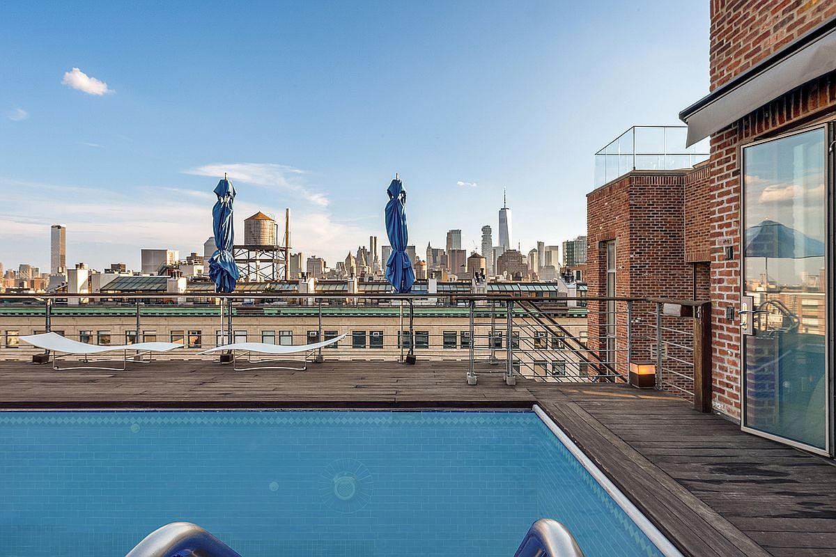 This stunning rooftop terrace features a sleek, rectangular swimming pool set into a rich wooden deck, offering an unobstructed, panoramic view of the New York City skyline. The space is furnished with minimalist white lounge chairs and closed blue umbrellas, creating a sophisticated urban oasis. The perspective captures the contrast between the warm, textured brick architecture of the building and the cool, expansive blue of the pool and sky.