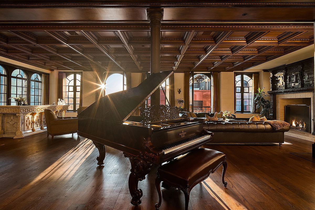 This opulent living room features a grand piano as its centerpiece, set against a backdrop of rich, dark wood coffered ceilings and polished hardwood floors. The space is elegantly furnished with a leather sofa, a stone fireplace, and a marble-topped bar, all bathed in the warm, golden glow of natural sunlight streaming through large arched windows. The overall atmosphere is one of sophisticated luxury and timeless architectural craftsmanship.