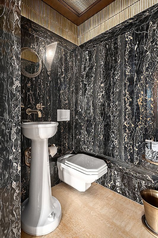 This luxurious powder room features dramatic black marble walls with striking gold veining, creating a sophisticated and opulent atmosphere. A sleek white pedestal sink and a wall-mounted toilet are complemented by warm, textured gold-toned ceiling accents and ambient wall lighting. The perspective is a wide-angle shot that captures the high-end materials and compact, elegant layout of this refined space.