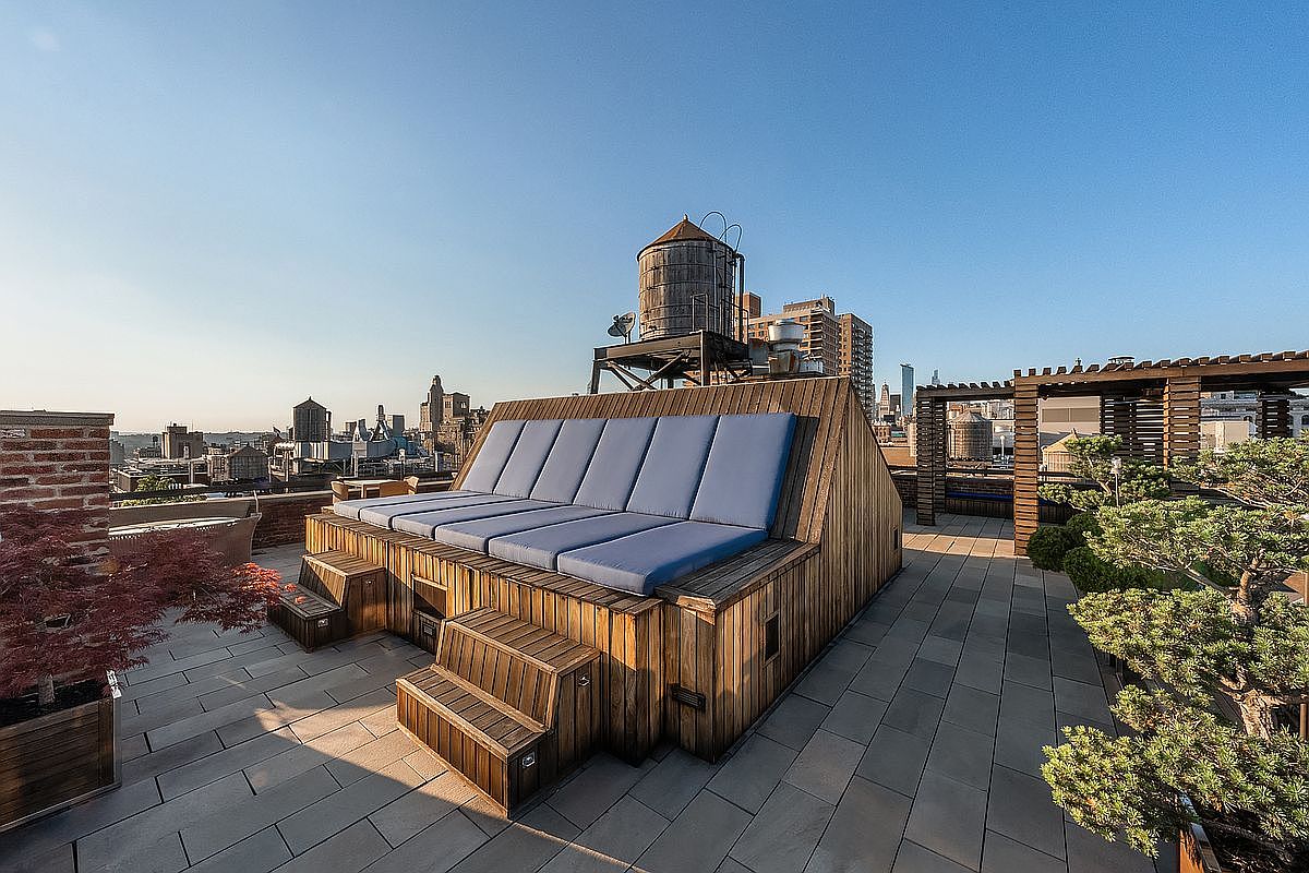 This expansive rooftop terrace features a custom-built wooden lounge structure with integrated blue cushions, providing a unique and luxurious seating area. The deck is paved with large, modern tiles and includes manicured bonsai-style trees, creating a serene urban oasis. The scene is captured from a slightly elevated perspective, showcasing the impressive city skyline and a classic water tower in the background.