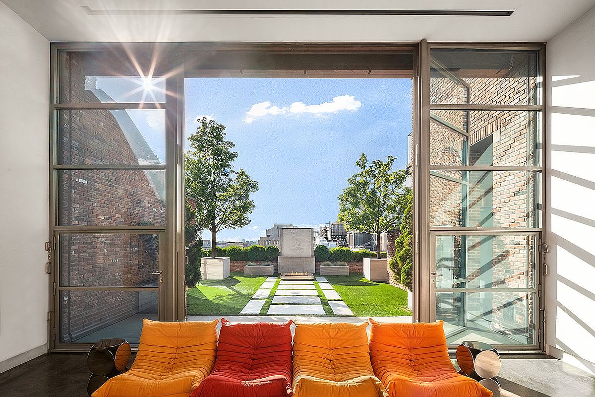 This sophisticated living room features a vibrant, modular orange and red sofa set positioned to face a massive, floor-to-ceiling glass wall that opens onto a private rooftop terrace. The terrace is beautifully landscaped with manicured trees, stone pavers, and a central fire feature, creating a seamless indoor-outdoor living experience. The contrast between the warm, plush interior seating and the crisp, modern architectural lines of the brick-walled terrace creates an inviting and luxurious urban retreat.