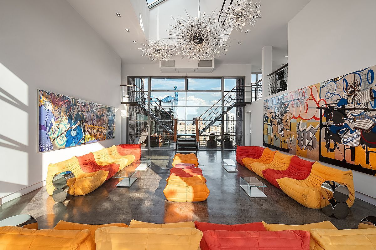 This expansive, high-ceilinged living room features a bold, contemporary aesthetic with vibrant orange and red modular seating arranged symmetrically. The space is anchored by large-scale, colorful graffiti-style murals on both side walls and illuminated by striking, starburst-inspired chandeliers. A massive floor-to-ceiling window wall at the rear provides an industrial-chic backdrop, showcasing a metal staircase and an urban view.