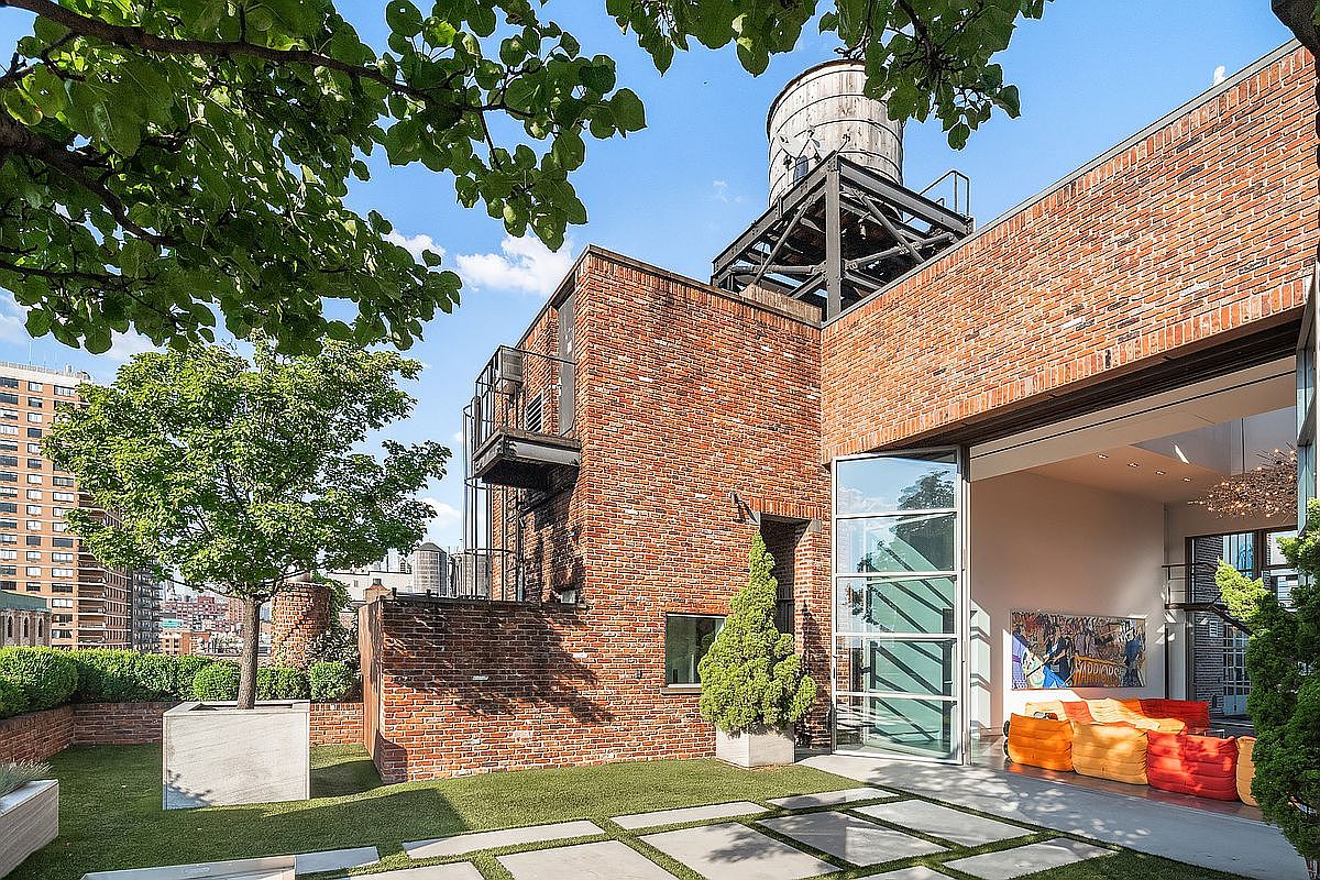 This stunning rooftop terrace features a blend of industrial brick architecture and modern landscaping, complete with a large water tower structure. The space includes a manicured lawn area with concrete pavers, a mature tree in a concrete planter, and a seamless transition into an indoor living area through expansive glass doors. The overall impression is one of sophisticated urban luxury, offering a private outdoor retreat with a view of the surrounding city skyline.