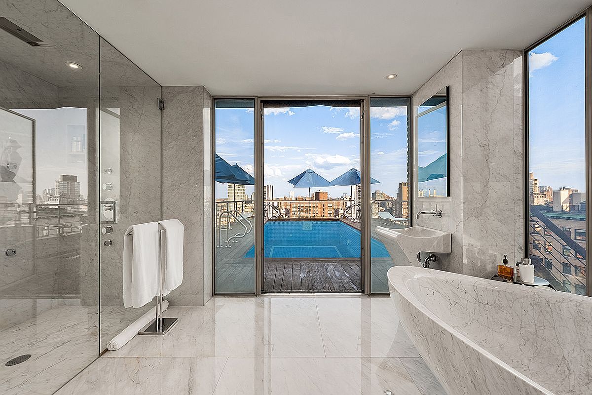 This luxurious primary bathroom features floor-to-ceiling marble tiling, a sleek freestanding soaking tub, and a spacious glass-enclosed walk-in shower. The room is anchored by a large floor-to-ceiling window that provides a seamless transition to a private outdoor terrace with a sparkling blue swimming pool and panoramic city views. The overall aesthetic is one of high-end sophistication, blending modern architectural design with an open, airy atmosphere.