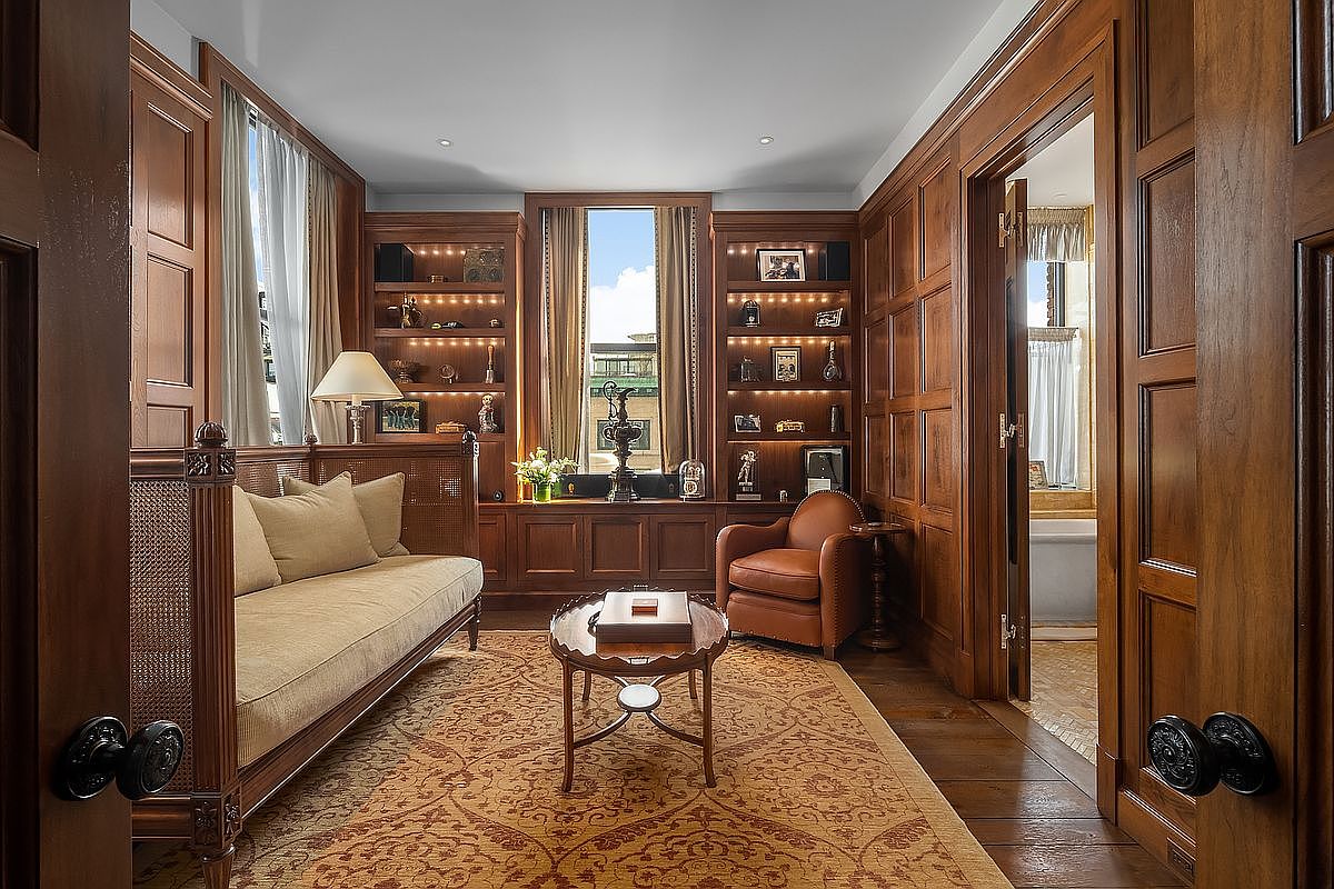 This sophisticated study exudes a classic, scholarly atmosphere with its rich, floor-to-ceiling wood paneling and built-in shelving units. The room is furnished with a plush, cane-sided sofa, a leather armchair, and a central wooden coffee table, all anchored by an ornate patterned rug. The perspective is framed through a doorway, looking into the warm, inviting space that features a glimpse of an adjacent bathroom.