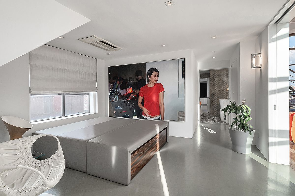 This modern, minimalist bedroom features a sleek, low-profile gray platform bed positioned against a large, striking photographic art piece. The room is characterized by polished concrete floors, a neutral color palette, and a contemporary white chair, creating a sophisticated and edgy urban aesthetic. Natural light streams in from the side window, highlighting the clean lines and open layout that leads into an adjacent hallway.