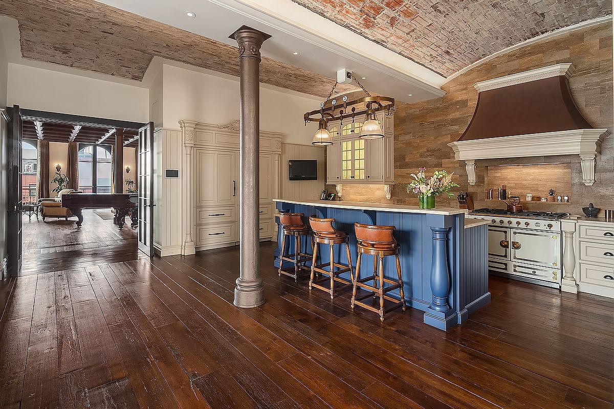 This sophisticated kitchen features a striking blend of rustic and industrial design, highlighted by a vaulted brick ceiling and a prominent dark metal support column. A deep blue kitchen island with leather-upholstered bar stools serves as the centerpiece, complemented by a professional-grade range and a custom-built range hood set against a textured stone wall. The perspective captures the open-concept flow from the kitchen into an adjacent living area, emphasizing the expansive dark hardwood flooring and high-end architectural character.