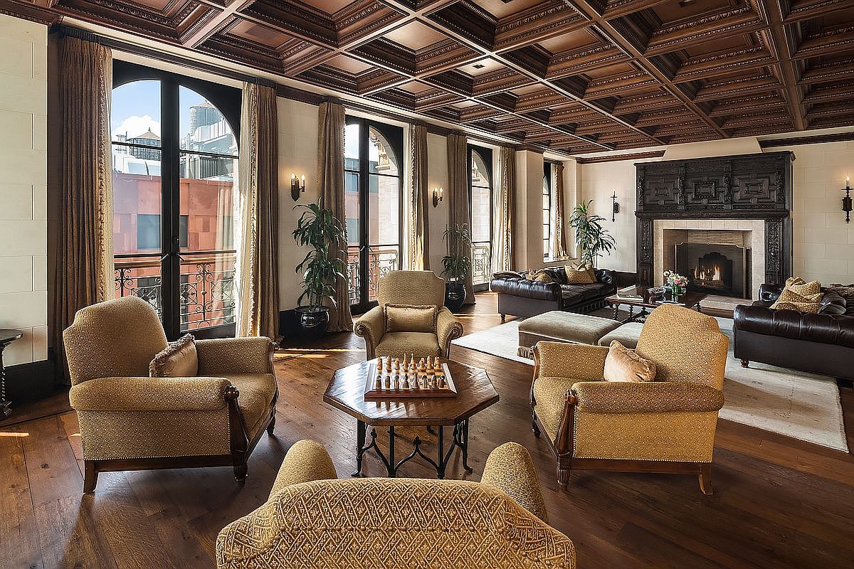 This grand, sophisticated living room features a stunning coffered wood ceiling and rich hardwood flooring, creating an atmosphere of timeless elegance. The space is anchored by a large, ornate dark wood fireplace and furnished with plush, textured armchairs arranged around a central chess table. Large arched windows with floor-to-ceiling drapery allow for abundant natural light and provide a view of the urban exterior, blending classic luxury with a comfortable, inviting layout.