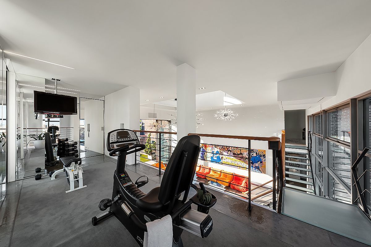 This modern, multi-level home gym features a sleek, industrial aesthetic with polished concrete floors and a prominent recumbent exercise bike in the foreground. The space overlooks a lower living area adorned with vibrant, artistic wall murals and orange seating, creating a dynamic and energetic atmosphere. Large windows and glass railings enhance the open-concept design, providing a bright and airy feel that seamlessly connects the workout area to the rest of the home.