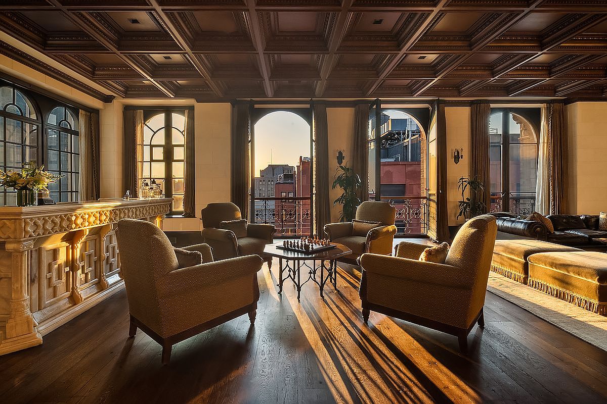 This sophisticated living room features a rich, coffered wood ceiling and dark hardwood floors, creating an atmosphere of timeless elegance. The space is anchored by a central seating area with four plush armchairs surrounding a chess table, all bathed in the warm, golden glow of a sunset streaming through arched balcony doors. A grand, intricately carved stone bar adds a touch of architectural grandeur, while the open layout seamlessly connects the refined interior to the urban cityscape beyond.