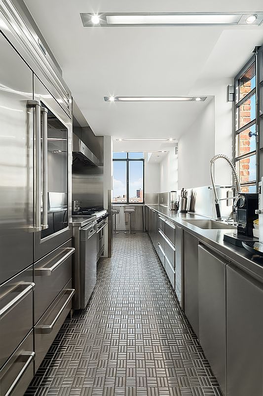 This sleek, galley-style kitchen features high-end stainless steel appliances, including a large refrigerator and a professional-grade range. The space is defined by its industrial aesthetic, highlighted by a unique metallic tiled floor and clean, minimalist cabinetry. A long countertop runs along the right side, leading the eye toward a bright window at the end of the room that offers an expansive city view.