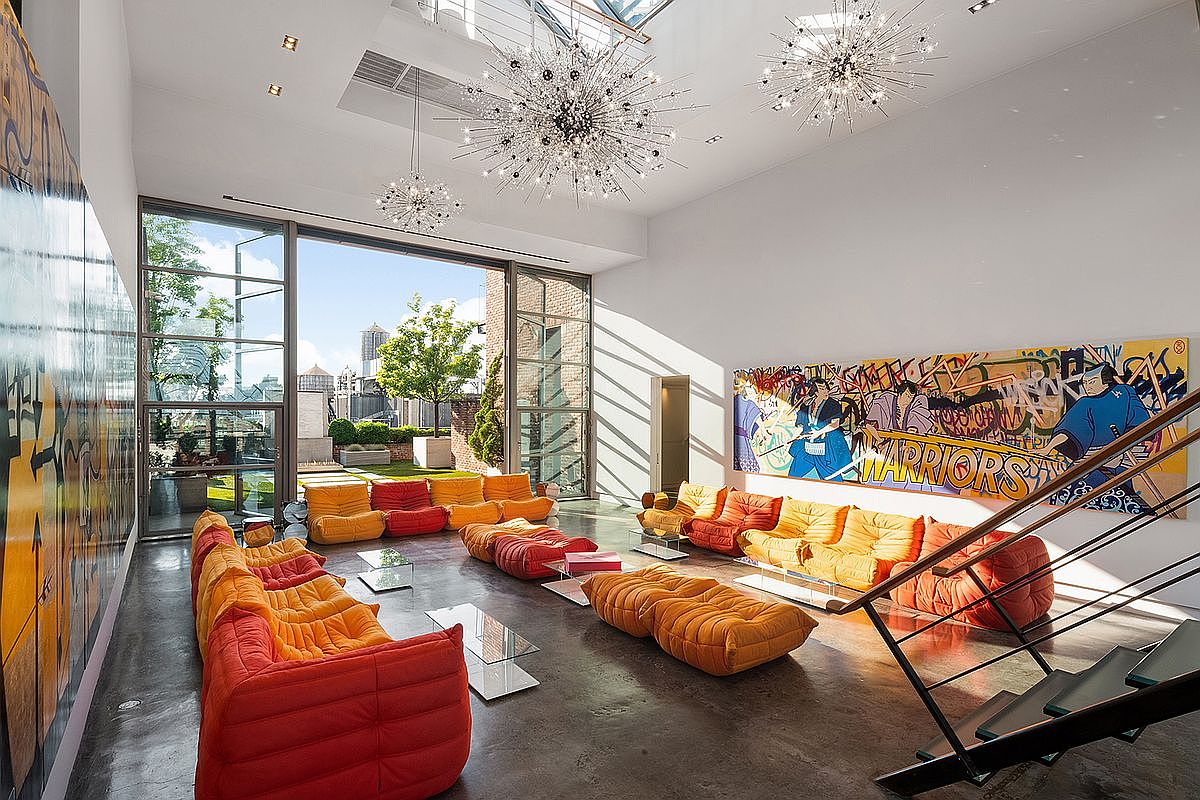 This expansive, high-ceilinged living room features a vibrant, modern aesthetic with modular, multi-colored seating arrangements in shades of orange and yellow. A large, striking mural depicting stylized warriors adorns the wall, while floor-to-ceiling glass doors open onto a private terrace, flooding the space with natural light. The room is accented by dramatic, starburst-style chandeliers and a sleek, industrial-style staircase, creating a sophisticated and artistic atmosphere.