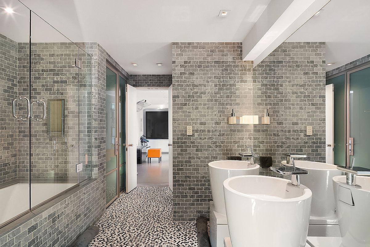 This modern primary bathroom features a striking aesthetic with grey brick-style wall tiles and a unique mosaic-tiled floor. The space is anchored by two sleek, white pedestal sinks and a glass-enclosed shower area, all reflected in a large, expansive mirror that enhances the room's depth. A doorway leads into an adjacent room, providing a sense of flow and connectivity within the home's layout.