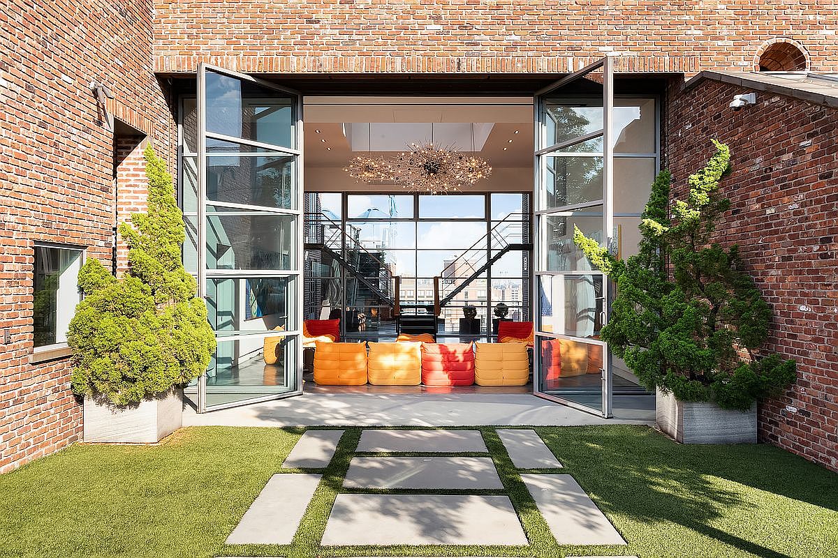 This image captures a sophisticated transition between a lush, manicured courtyard and a modern, industrial-style interior living space. The foreground features a geometric arrangement of concrete pavers set into vibrant green turf, flanked by two large, potted evergreen trees. The massive, floor-to-ceiling glass doors are swung wide open, revealing a bright, contemporary living area furnished with colorful, modular seating and an impressive, starburst-style chandelier.