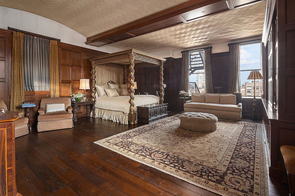 This opulent primary bedroom features rich, dark wood-paneled walls and a dramatic four-poster gold-leaf bed as its centerpiece. The room is anchored by a large, intricate area rug and a tufted ottoman, while a sophisticated coffered ceiling adds architectural depth. Large windows draped in heavy curtains allow natural light to illuminate the classic, luxurious decor.