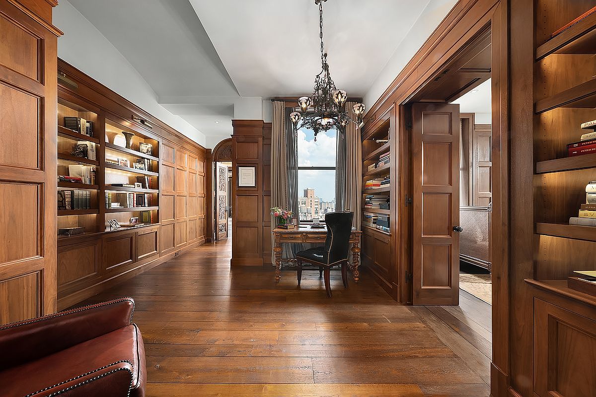 This sophisticated home office exudes a classic, scholarly atmosphere with its rich, floor-to-ceiling wood paneling and extensive built-in bookshelves. A central desk is positioned in front of a window that provides a view of the city skyline, while an ornate chandelier hangs above, adding a touch of elegance. The warm wood tones and leather furniture create a timeless, luxurious, and studious environment.