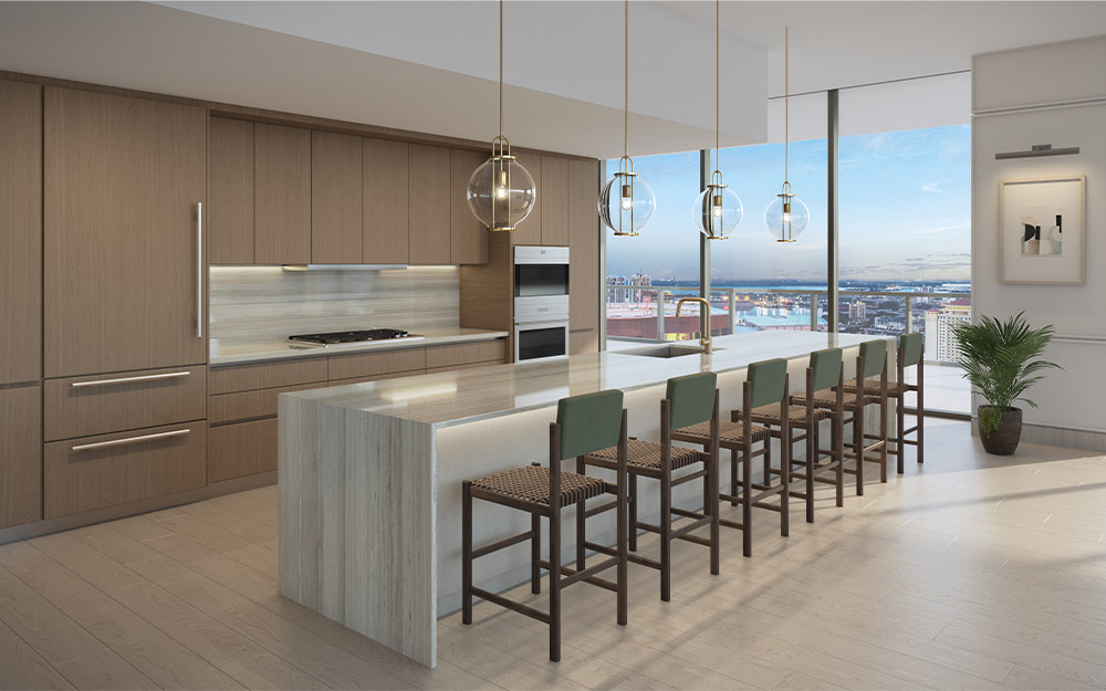 This is a modern kitchen featuring light wood cabinetry and a large island with a waterfall countertop. The kitchen is equipped with stainless steel appliances and pendant lighting. Large windows offer a panoramic city view, enhancing the luxurious feel of the space.