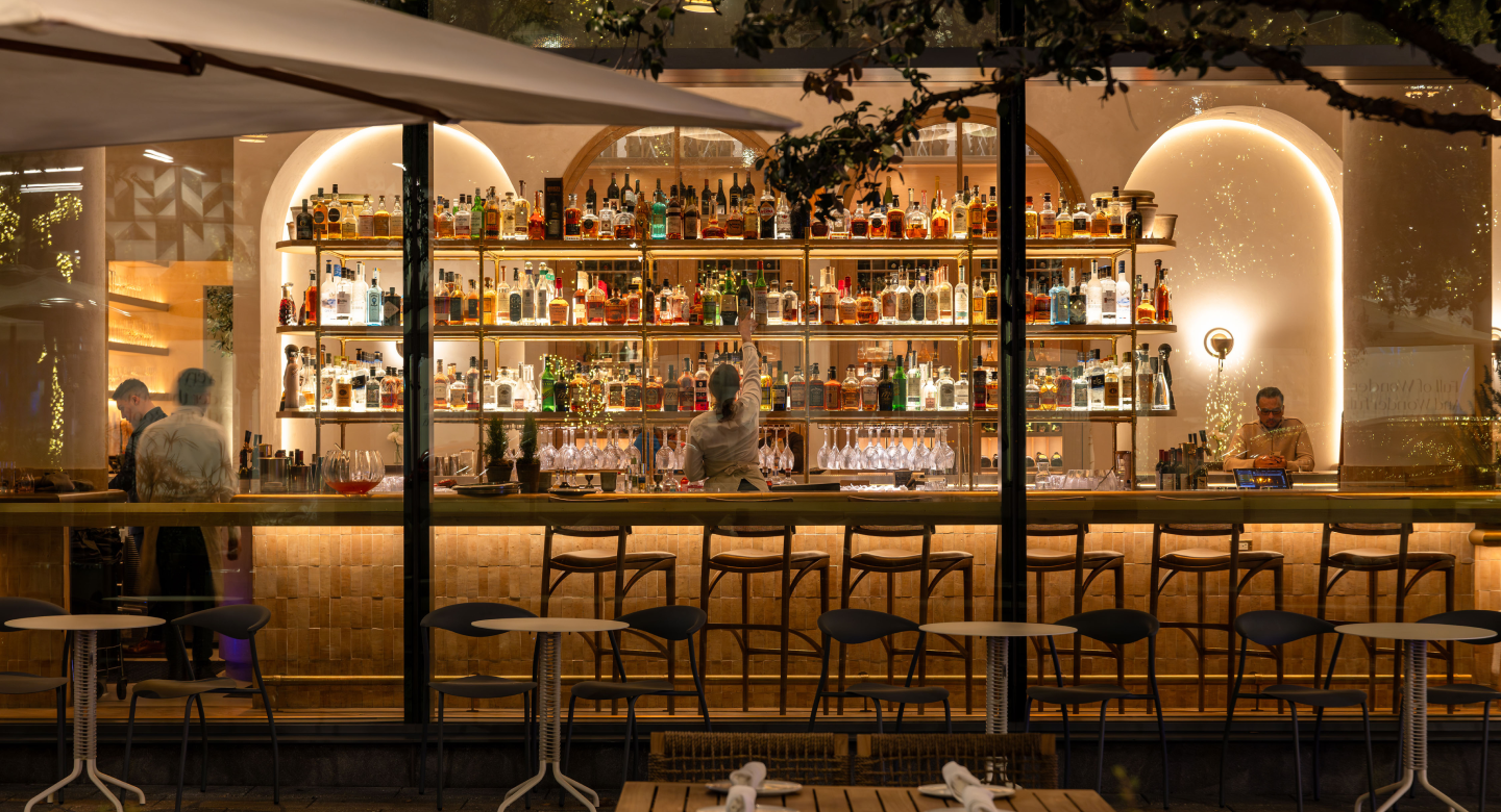This image showcases the interior of a restaurant or bar, featuring a well-stocked bar with numerous bottles displayed on shelves. The foreground includes tables and chairs, suggesting a dining area. The warm lighting and stylish decor create an inviting and upscale atmosphere, perfect for attracting potential customers.