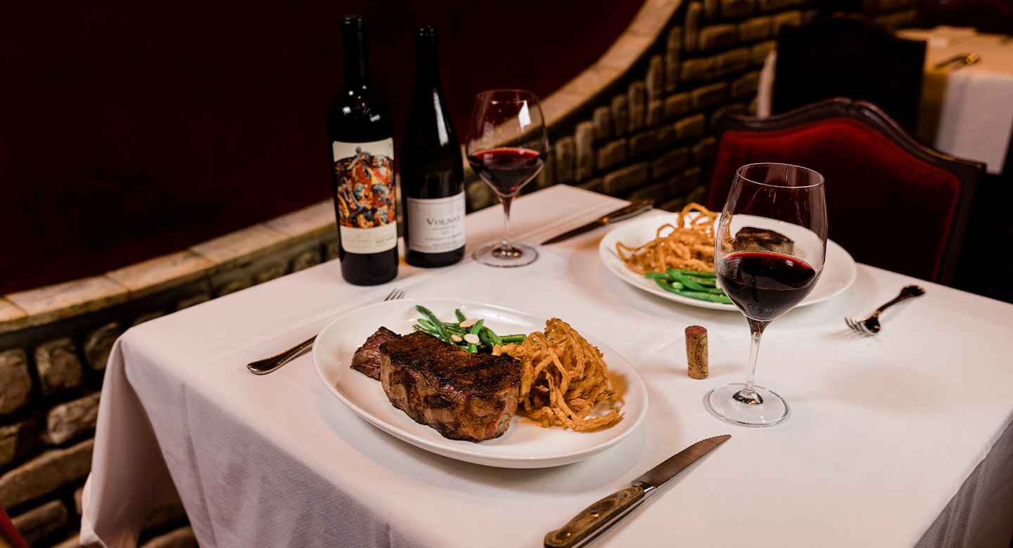 This image showcases a fine dining experience, featuring two plates of steak with green beans and onion strings, accompanied by two bottles of red wine and wine glasses filled with red wine. The setting is elegant, with a white tablecloth and a dimly lit background, suggesting a high-end restaurant or a sophisticated home dining room. The focus is on the culinary presentation and the ambiance of a luxurious meal.