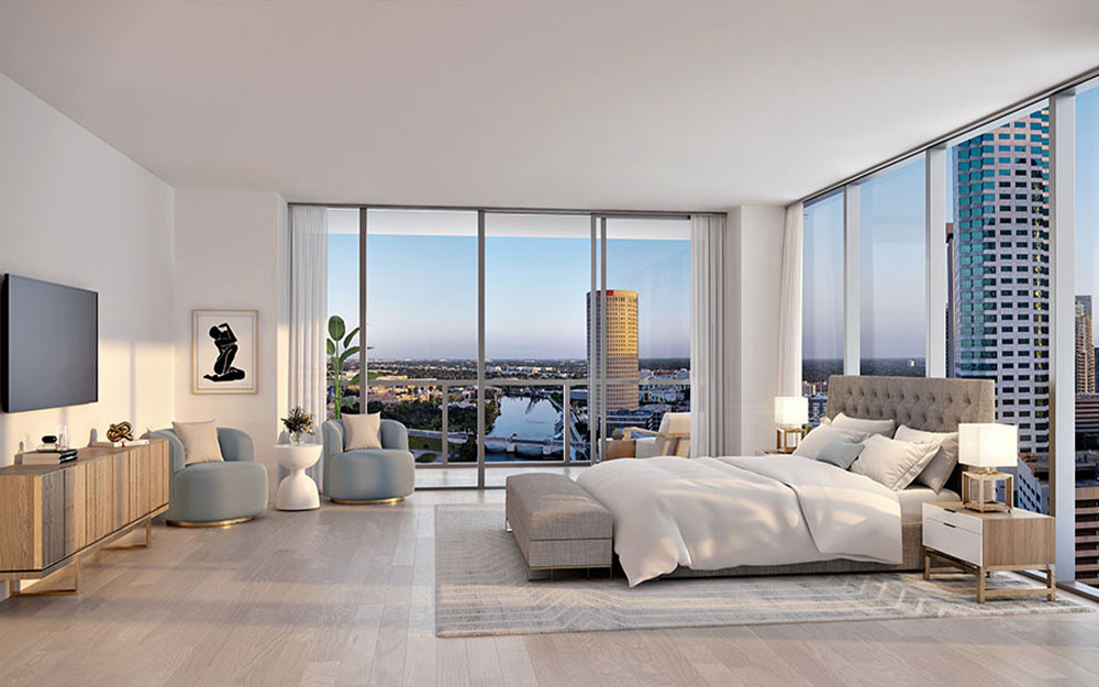 This is a luxurious primary bedroom with floor-to-ceiling windows offering a stunning city view. The room features a king-size bed with a tufted headboard, light-colored bedding, and modern nightstands with lamps. A console table with a TV and artwork adds to the room's elegance, complemented by two stylish armchairs.