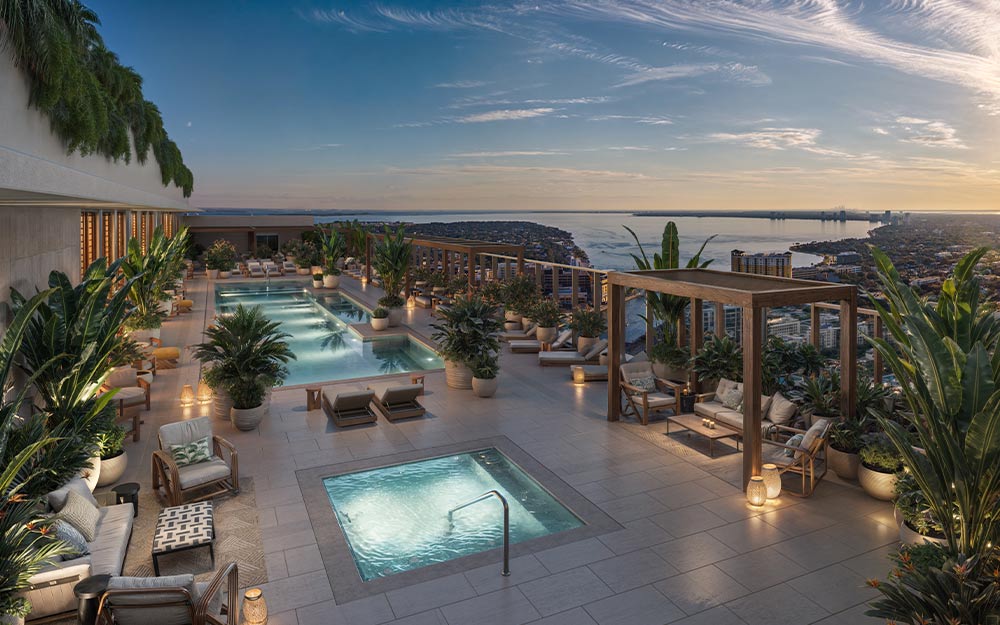 This image showcases a luxurious rooftop pool and spa area, complete with lounge chairs, lush greenery, and wooden pergolas. The pools are illuminated, creating a serene and inviting atmosphere. The view extends to a distant cityscape and body of water, enhancing the property's appeal.