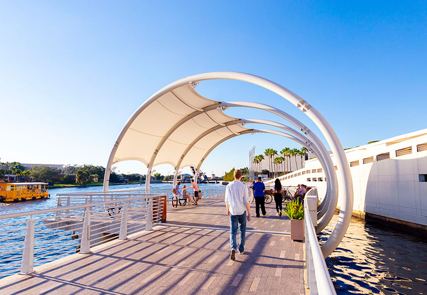 This image showcases a modern waterfront walkway featuring a unique arched canopy structure providing shade. People are seen strolling and enjoying the waterfront view, with a yellow water taxi visible in the background. The scene evokes a sense of community and leisure, highlighting the amenity's appeal.