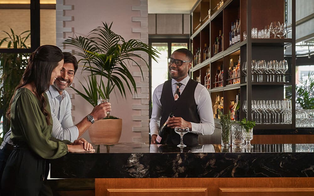 This interior shot showcases a stylish bar area, featuring a dark marble countertop and a well-stocked shelving unit with various bottles and glassware. A smiling bartender serves a couple, creating a welcoming and upscale atmosphere. The space is well-lit and decorated with plants, adding a touch of freshness.