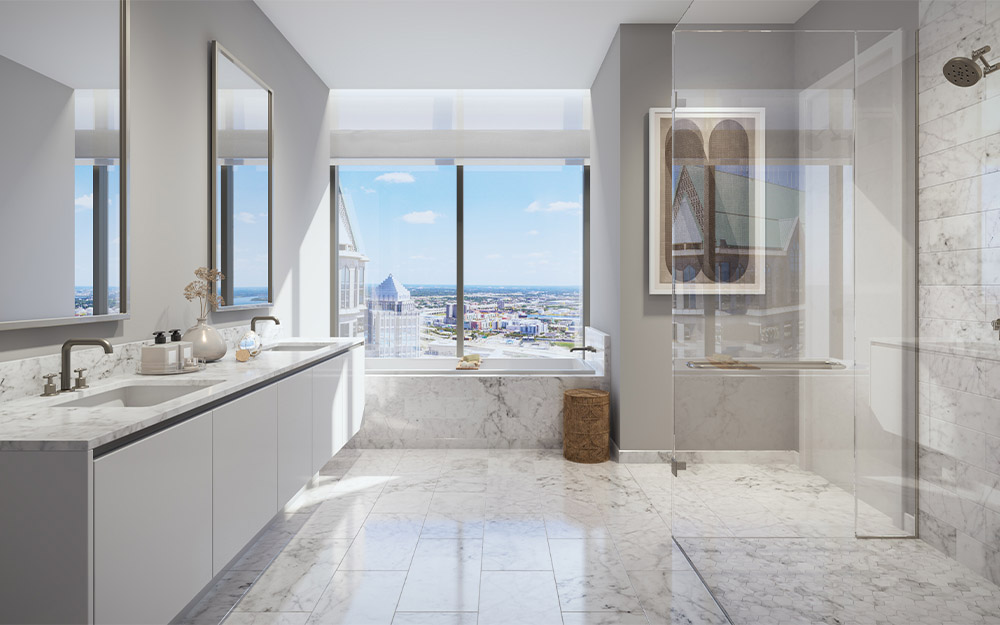 This is a luxurious primary bathroom featuring a double vanity with marble countertops and sleek white cabinetry. A large window offers a stunning city view, while a freestanding bathtub sits beneath it. The bathroom also includes a glass-enclosed shower and marble flooring, creating a spa-like atmosphere.