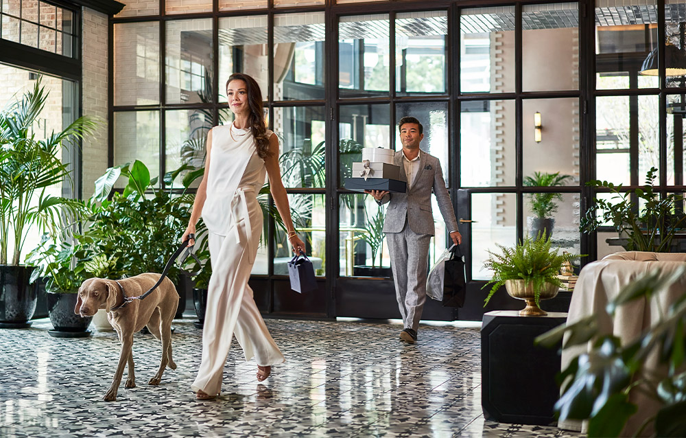 This interior shot showcases a stylish hallway with a patterned tile floor, large windows, and potted plants, creating a bright and inviting atmosphere. A woman walks a dog while a man carries gifts, suggesting a luxurious residential or hotel setting. The space is well-lit and features modern architectural details, enhancing its appeal.