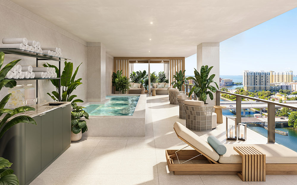 This image showcases a luxurious outdoor patio/balcony area featuring a small pool or spa, lounge chairs, and comfortable seating. The space is adorned with lush greenery, creating a serene and private atmosphere. The view overlooks a cityscape and waterway, adding to the upscale ambiance and highlighting the property's prime location.