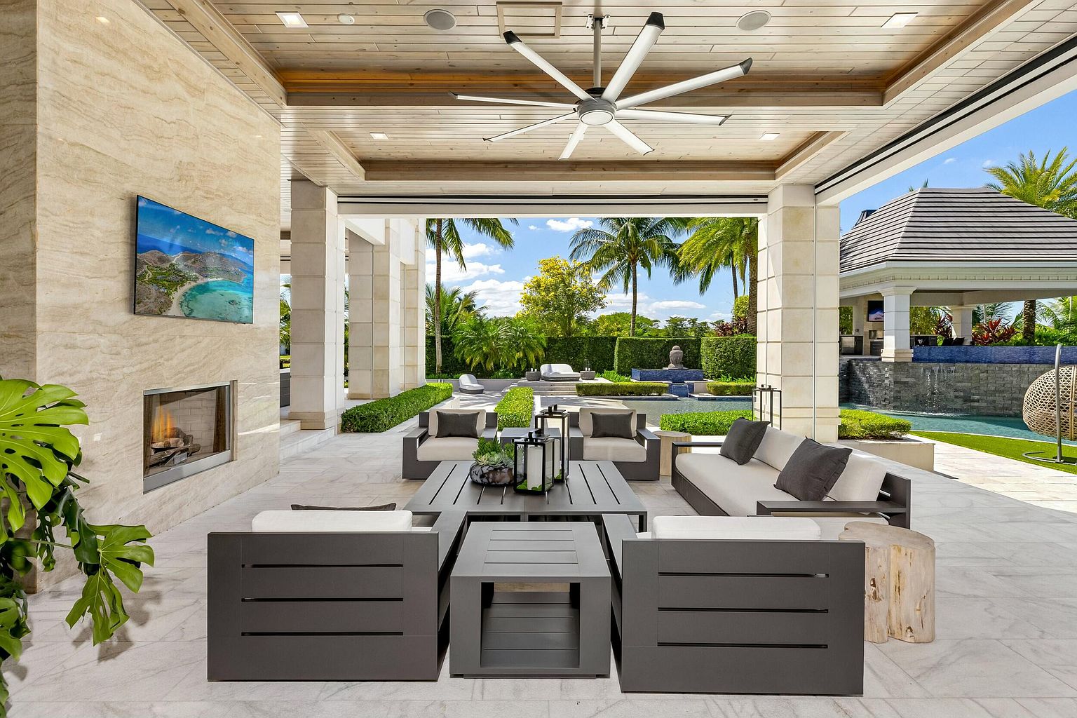 This image showcases a luxurious outdoor patio area, featuring modern dark gray furniture with light cushions arranged around a central coffee table. The patio is covered by a wooden-paneled ceiling with a large ceiling fan, and a stone fireplace with a mounted TV adds a touch of indoor comfort. The space opens up to a beautifully landscaped garden and pool area, creating a seamless transition between indoor and outdoor living.